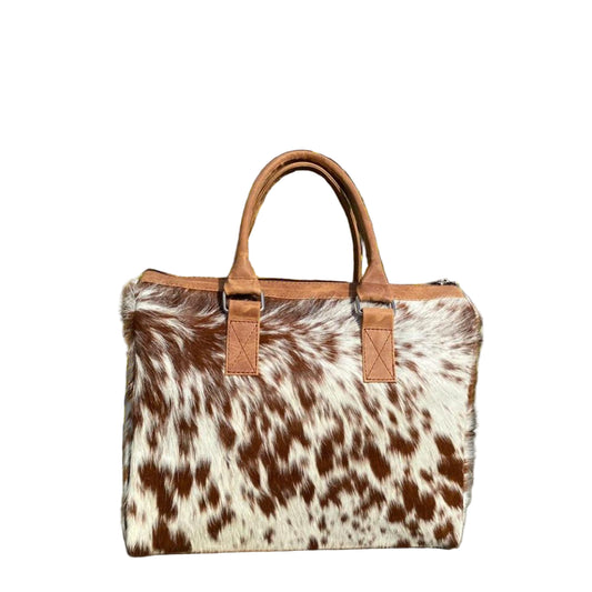 Brown and white cowhide bag with brown leather handles against a green plant background