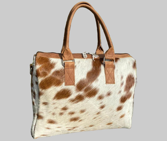 Brown and white cowhide leather handbag with brown leather handles against a green plant background.